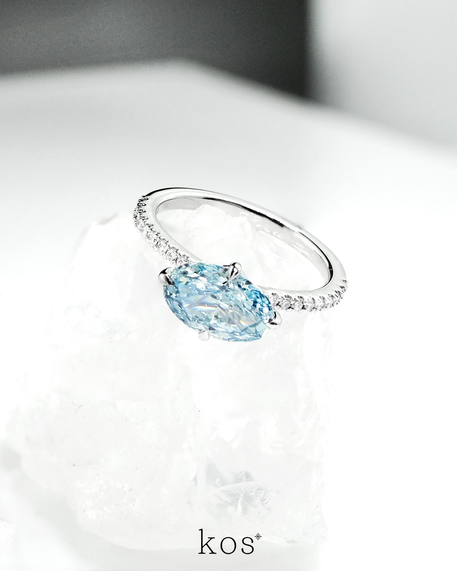 แหวนหมั้นเพชร Oval สีฟ้าทรง วางแนวนอน (East-West) บนตัวเรือนทองคำขาวฝังเพชร ดีไซน์แบรนด์ kos
